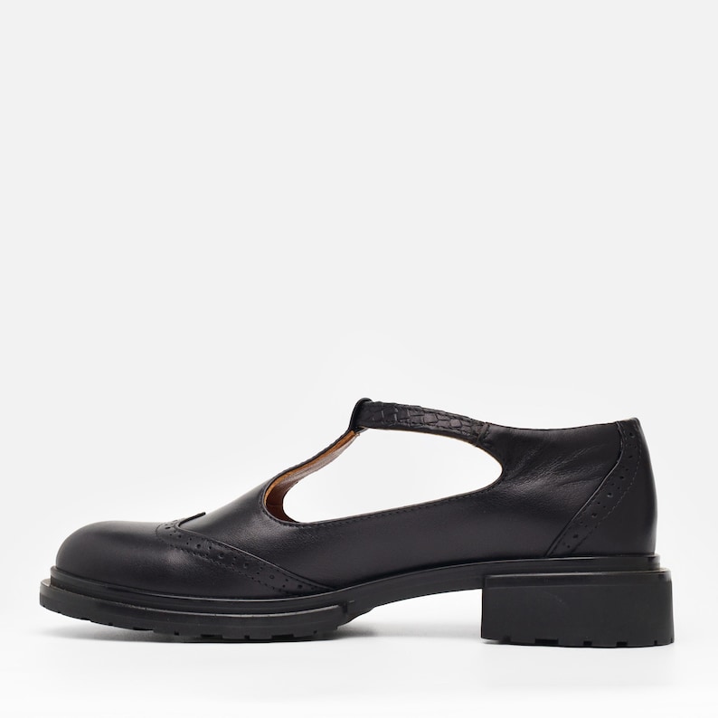 May include: A black leather T-strap shoe featuring a rounded toe, decorative brogue perforations along the vamp, and a textured strap. The shoe is constructed with a low, block heel and a thick, lugged rubber sole for a sturdy, flat profile.