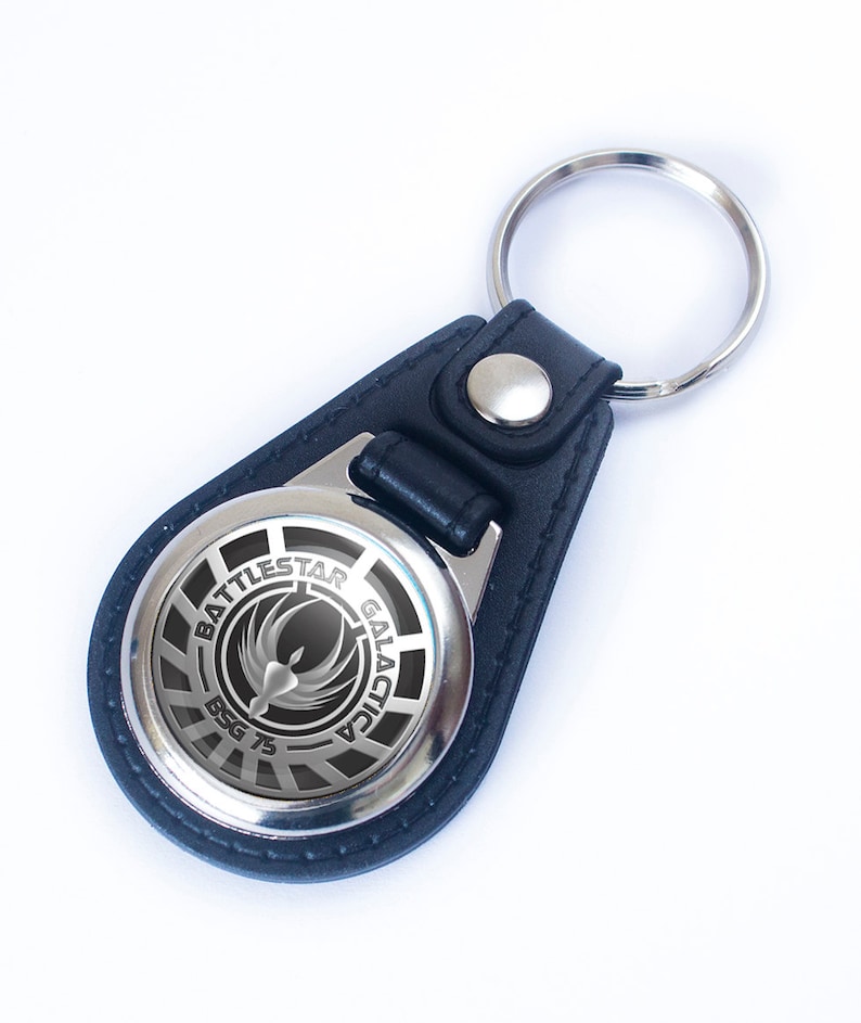 May include: A black leather keychain with a silver circular charm featuring the Battlestar Galactica logo and the text "Battlestar Galactica" and "BSG 75".