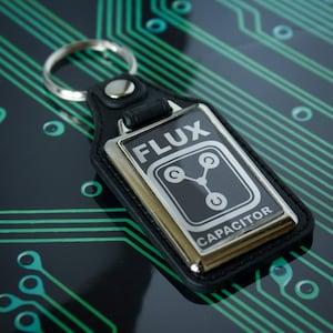 May include: Black leather keychain with a silver metal charm. The charm has the words "Flux Capacitor" and a stylised image of a circuit board.