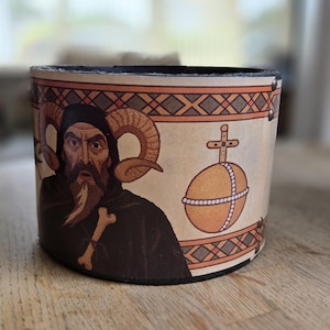 May include: A cylindrical leather container featuring an illustration of a robed figure with ram horns, a long beard, and a cross. The container has a black interior and is decorated with an orange and brown geometric pattern.