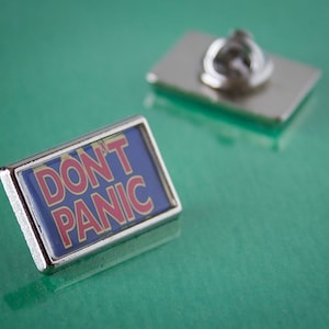 May include: A silver pin with a blue background and the words "DON'T PANIC" in red and white letters.