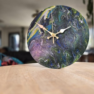 May include: Circular wall clock featuring an abstract design in blue, green, yellow, and purple. The clock has gold-coloured hands and a gold-coloured centre. The clock is displayed on a wooden surface.