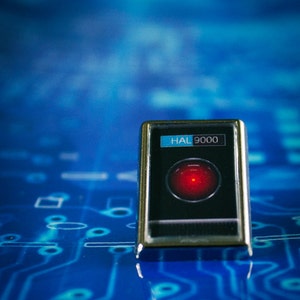 May include: A silver pin with a black background and a red circle in the centre. The text "HAL 9000" is displayed on the pin.