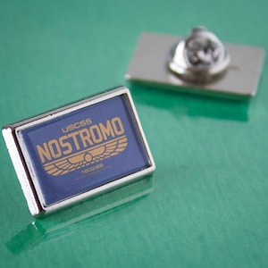 May include: A silver pin with a blue background and gold lettering that reads "USCSS NOSTROMO" with a gold winged symbol and the text "180288" underneath.