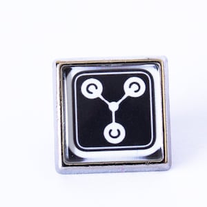 May include: A silver-toned pin featuring a black and white image of the iconic "flux capacitor" from the Back to the Future film franchise.