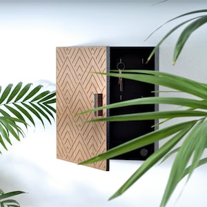 Könnte beinhalten: Ein hölzerner Schlüsselhalter mit geometrischem Design auf der Fronttür. Der Schlüsselhalter ist geöffnet und zeigt einen schwarzen Innenraum mit mehreren Haken für Schlüssel. Ein kleiner brauner Griff ist sichtbar. Er ist an einer weißen Wand montiert.