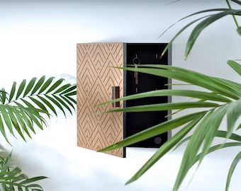 May include: A wooden key holder with a geometric design on the front door. The key holder is open, revealing a black interior with several hooks for keys. A small brown handle is visible. The key holder is mounted on a white wall.