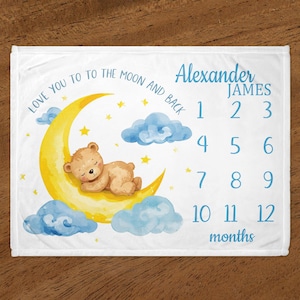 May include: White baby milestone blanket with a watercolor illustration of a sleeping teddy bear on a yellow crescent moon, surrounded by blue clouds and stars. Text includes "Love you to the moon and back," a name, and numbers 1-12 for months.
