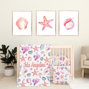 May include: A white crib bedding set with a pink, blue, and coral seashell and starfish pattern. The set includes a blanket, a pillow, and three wall prints. The blanket and pillow feature the text "Isla Angeline".