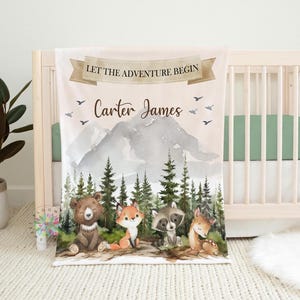 May include: A personalized baby blanket with a woodland theme. The blanket features a mountain range, a forest, and a variety of animals, including a bear, a fox, a raccoon, and a deer. The blanket is decorated with the text "Let the adventure begin" and the name "Carter James".