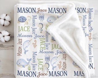 May include: A white baby blanket with a marine life design. The blanket features illustrations of a turtle, jellyfish, whale, seahorse, and narwhal. The names "MASON" and "JACE" are printed in various fonts and colors across the blanket.