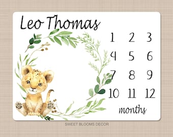 May include: A white baby milestone card with a green leafy wreath and a cute cartoon lion cub. The card has the name "Leo Thomas" and numbers 1 through 12 for each month of the baby's first year.