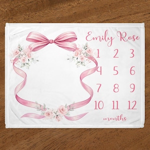 May include: A white baby milestone blanket with a pink bow, floral design, and the text "Emily Rose" and "months" along with numbers 1 through 12.