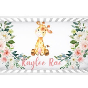 May include: A white crib sheet with a pink floral border and a watercolor illustration of a giraffe in the center. The name "Kaylee Rae" is written in pink script below the giraffe.