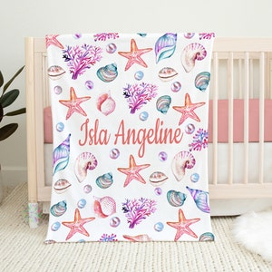 May include: A white baby blanket with a pink, coral, and blue seashell and starfish pattern. The blanket has the name "Isla Angeline" written in pink on it.