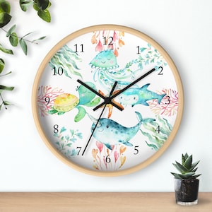 May include: A white round clock with a wooden frame features a watercolor design of sea creatures, including a jellyfish, a turtle, a shark, and a narwhal. The clock face has black numbers and hands.