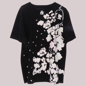 May include: A black short-sleeved t-shirt featuring a graphic print of a cherry blossom branch extending from the top right shoulder down to the bottom hem, with scattered pink and white flower petals across the front and a small ship logo inside.