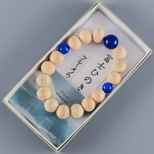 May include: A bracelet made of light-colored wooden beads and three smooth, round, dark blue beads. The bracelet is presented in a rectangular box with a card featuring the text Fuji Hinoki and Japanese characters, along with a blue-toned image.