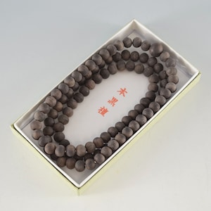 May include: A necklace or prayer beads made of dark brown wooden beads, presented in a rectangular box. The beads are smooth and spherical. The box has a white interior and a light green exterior. Japanese text is visible on the card inside the box.