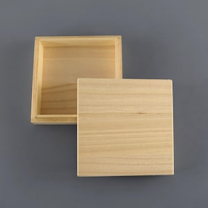 Könnte beinhalten: Eine kleine, offene Holzkiste mit separatem Deckel. Die Kiste und der Deckel sind aus hellem Holz gefertigt, mit sichtbaren Maserungsmustern. Die Kiste ist quadratisch geformt, und der Deckel ist etwas größer und liegt neben der offenen Kiste.