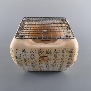 May include: A beige ceramic hibachi grill with a metal grate. The grill has Japanese characters written on the side. The grill is designed for cooking and grilling food.
