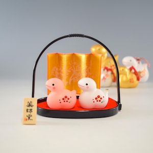 以下が含まれることがあります： 赤い花柄の小さな陶器製の鴨の置物2体。鴨は黒と赤のトレイの上に座っており、トレイには黒い取っ手がついています。トレイは金の屏風の前にあります。