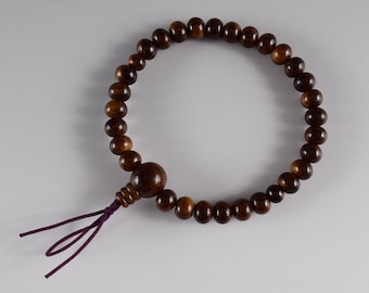 May include: A circular bracelet composed of polished dark brown wooden beads of uniform size, featuring one larger central bead and a smaller ribbed bead attached to a thin purple cord that hangs down from the main circular strand.