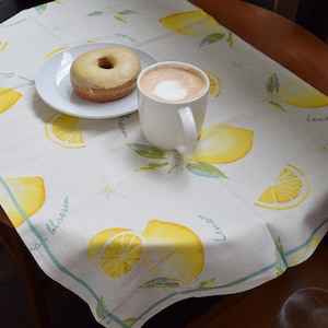 Op de afbeelding: Een witte keramische mok gevuld met koffie en een geglazuurde donut op een wit bord. Het bord en de mok staan op een wit tafelkleed met een citroenprint.
