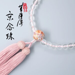 May include: A clear crystal prayer bead necklace with a pink tassel and a decorative bead with floral designs. The necklace has a red stamp with Japanese characters.