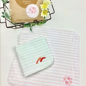 May include: Two square cotton hand towels with soft stripes. One towel is light green with a small embroidered shrimp sushi piece. The other is light pink with a cluster of embroidered pink cherry blossoms in the corner. Includes a paper tag.