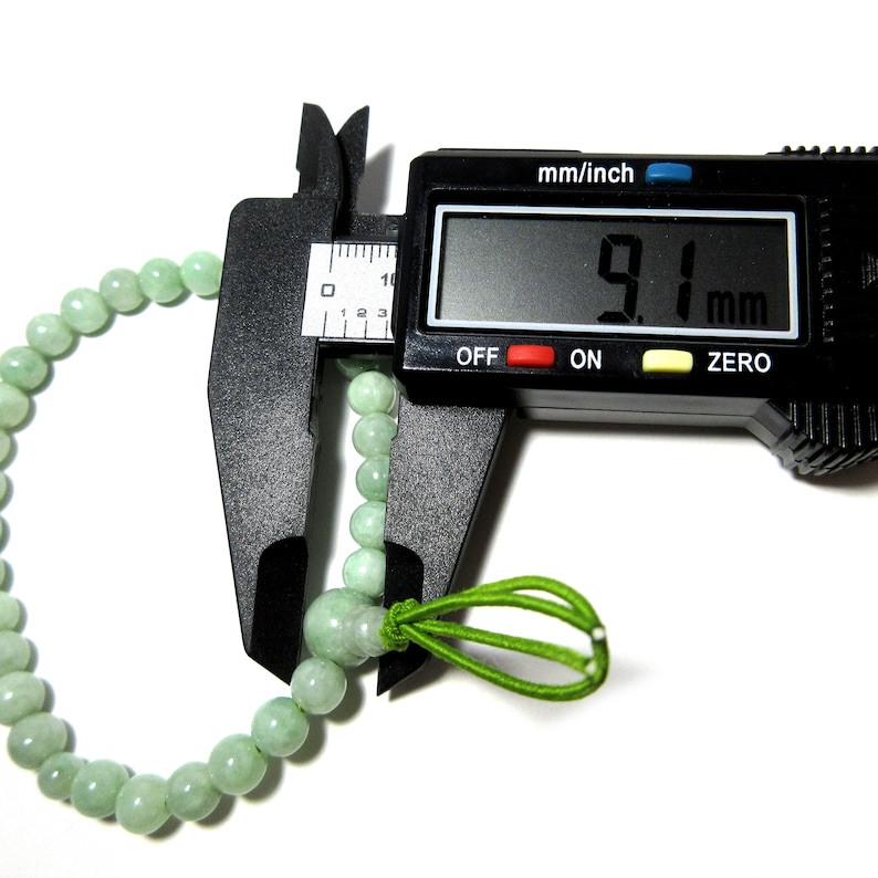 May include: A green jade bracelet with a green elastic cord closure. The bracelet is being measured with a digital caliper, which reads 9.1 mm.