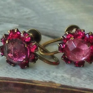 May include: A pair of vintage clip-on earrings with a large, faceted, pink rhinestone surrounded by smaller, red rhinestones. The earrings have a gold-tone metal setting.
