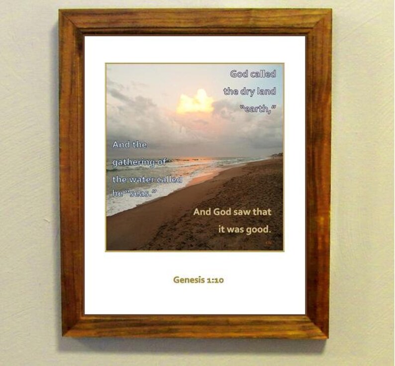 May include: A framed print with a beach scene at sunset. The print features a Bible verse from Genesis 1:10, which reads: "And the gathering of the water called he "seas." God called the dry land "earth," And God saw that it was good. Genesis 1:10"