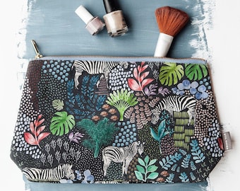 Könnte beinhalten: Eine schwarze Kosmetiktasche mit Reißverschluss und einem bunten Druck von Zebras und tropischen Pflanzen. Die Tasche hat einen goldenen Reißverschluss und ein blaues Futter. Die Tasche liegt auf einer blauen Oberfläche mit zwei Flaschen Nagellack und einem Make-up-Pinsel.