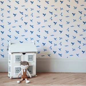 May include: A white doll's house with a doll inside. The dollhouse has two doors and two windows. The dollhouse is sitting on a wooden floor. The wall behind the dollhouse has a blue and white wallpaper with a pattern of flying birds.