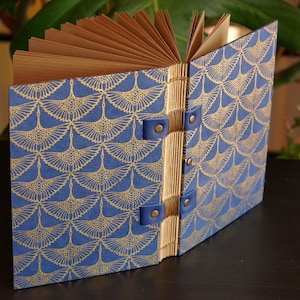 Könnte beinhalten: Offenes Notizbuch mit blauem Einband und goldenem Fächer-Muster. Die Seiten sind hellbraun, und der Rücken ist mit blauen Lederriemen und Messingnieten gebunden. Das Notizbuch ist geöffnet und zeigt die Seiten.