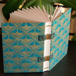 Peut inclure: Un journal ouvert avec une couverture à motifs turquoise et dorés. Le motif de la couverture présente un motif répétitif en forme d'éventail. Le journal est relié avec du fil beige et sécurisé par des lanières en cuir turquoise. Les pages sont blanches.