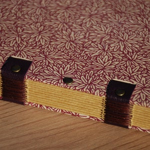 Peut inclure: Un journal de couleur bordeaux et crème avec un motif floral sur la couverture. Le journal a une reliure en cuir marron avec des coutures décoratives et des accents en laiton.