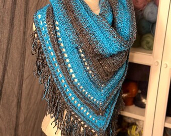 May include: A handmade, blue and gray crocheted shawl with fringe. The shawl is displayed on a mannequin and features a striped pattern with openwork detailing. The fringe is a darker shade of gray. The shawl is a triangular shape.