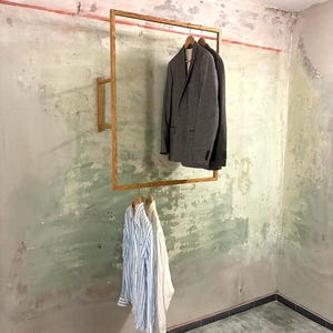 Könnte beinhalten: Ein hölzerner Kleiderständer an der Wand, der einen grauen Blazer und einen dunklen Blazer auf der oberen Stange sowie zwei Hemden mit Knöpfen auf der unteren Stange hält. Die Hemden sind blau-weiß gestreift und weiß. Die Wand ist in Grün- und Grautönen gestrichen.