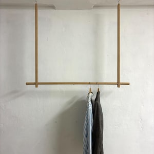 Könnte beinhalten: Eine hängende Kleiderstange aus Holz. Die Stange ist an der Decke befestigt und hat eine horizontale Stange mit zwei vertikalen Stützen. Ein gestreiftes Hemd und eine dunkelgraue Jacke hängen daran.