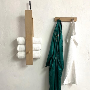 May include: A vertical light wood towel rack holding three folded white towels, and a horizontal light wood wall hook rack holding a dark green satin robe and a white towel. The wood fixtures feature a natural grain and simple, clean lines.