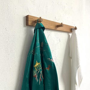 May include: A horizontal wooden wall rack with three cylindrical pegs. A green satin robe with embroidered pagoda and floral motifs hangs from the left peg, while a plain white towel hangs from the right peg against a textured white wall.