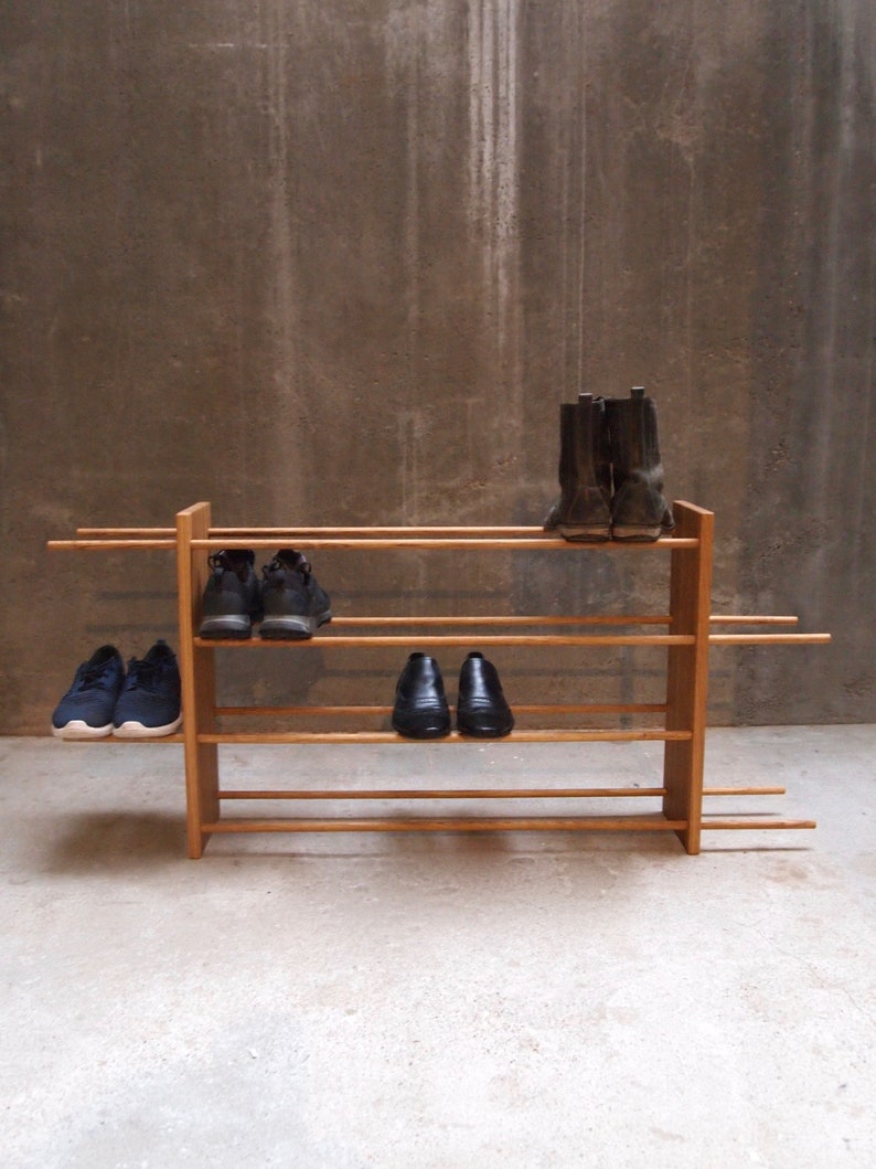 Op de afbeelding: Een houten schoenenrek met vier planken, met daarop een paar zwarte laarzen, twee paar zwarte schoenen en een paar zwarte sneakers. Het rek staat op een betonnen vloer.