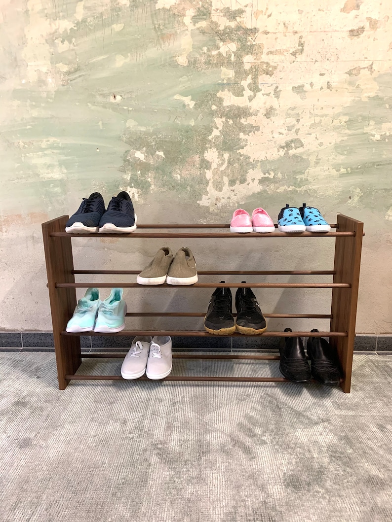 Op de afbeelding: Een houten schoenenrek met vier planken, met daarop verschillende schoenen, waaronder sneakers, sandalen en laarzen. Het rek staat op een grijs tapijt.