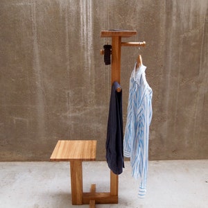 May include: A wooden coat rack with a shelf and a small seat. The rack has a single hook for hanging clothes and a small shelf for storing items. The seat is made of wood and is attached to the rack with a metal bracket.