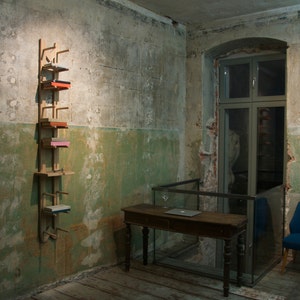 May include: A wooden shelf unit with five shelves, each holding a different colored box. The shelf unit is mounted on a wall in a room with a wooden table and a blue chair.