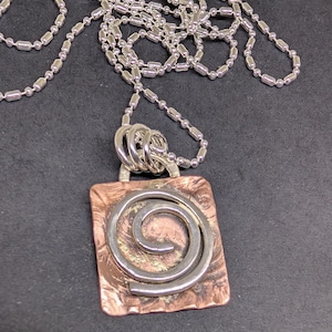 May include: A silver-toned necklace with a square pendant featuring a copper-colored background and a silver spiral design. The necklace has a ball chain and a silver clasp. The pendant is approximately 1.5 inches square.