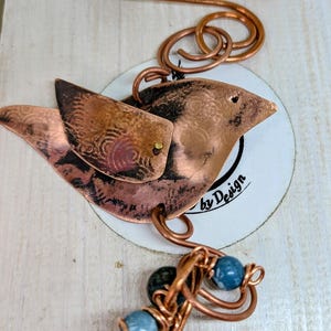 May include: A handcrafted copper bird ornament with etched details and a beaded accent. The bird has a layered wing design and is attached to a copper wire loop. The ornament includes a cluster of blue and green beads. The text "by Design" is visible.