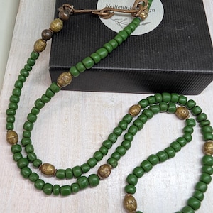 May include: A long necklace featuring small, round, matte green beads interspersed with larger, textured, metallic gold-toned beads. The necklace includes a copper-colored metal clasp mechanism with a hook and loop closure at the top.